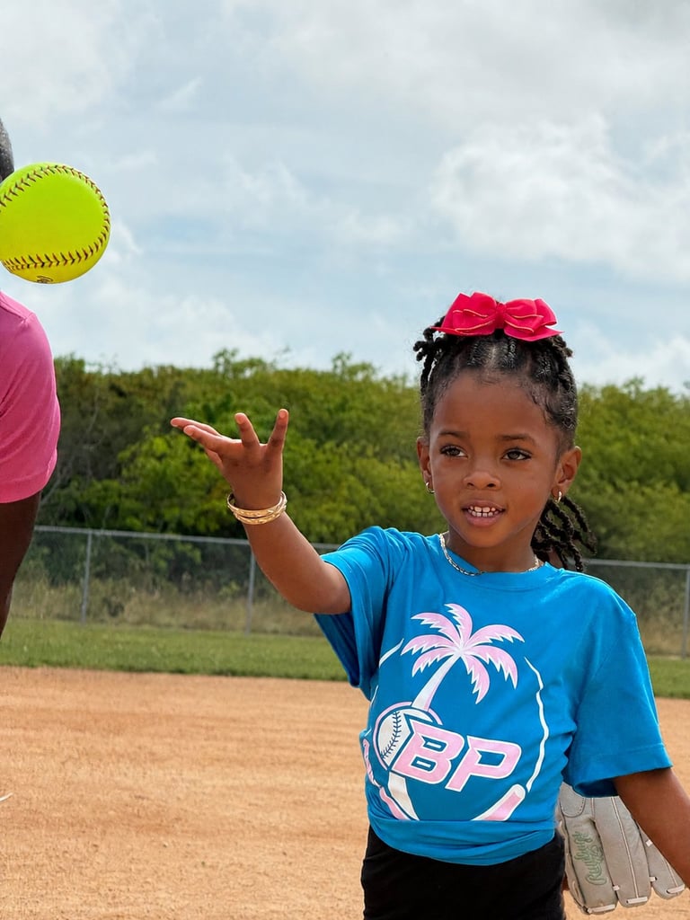 Young girl in blue shirt with pink bow catching yellow softball on dirt baseball field