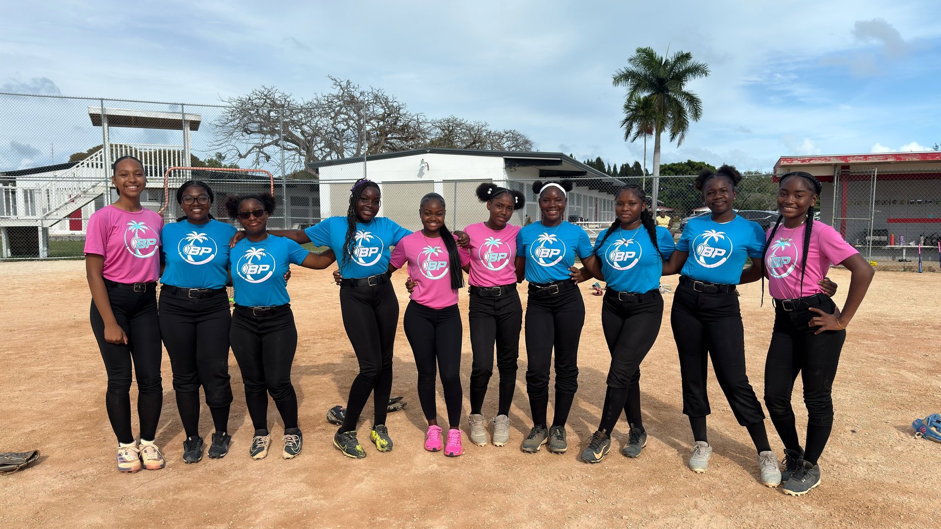 Group of young women wearing pink and blue volunteer shirts standing together in a line on a dirt field